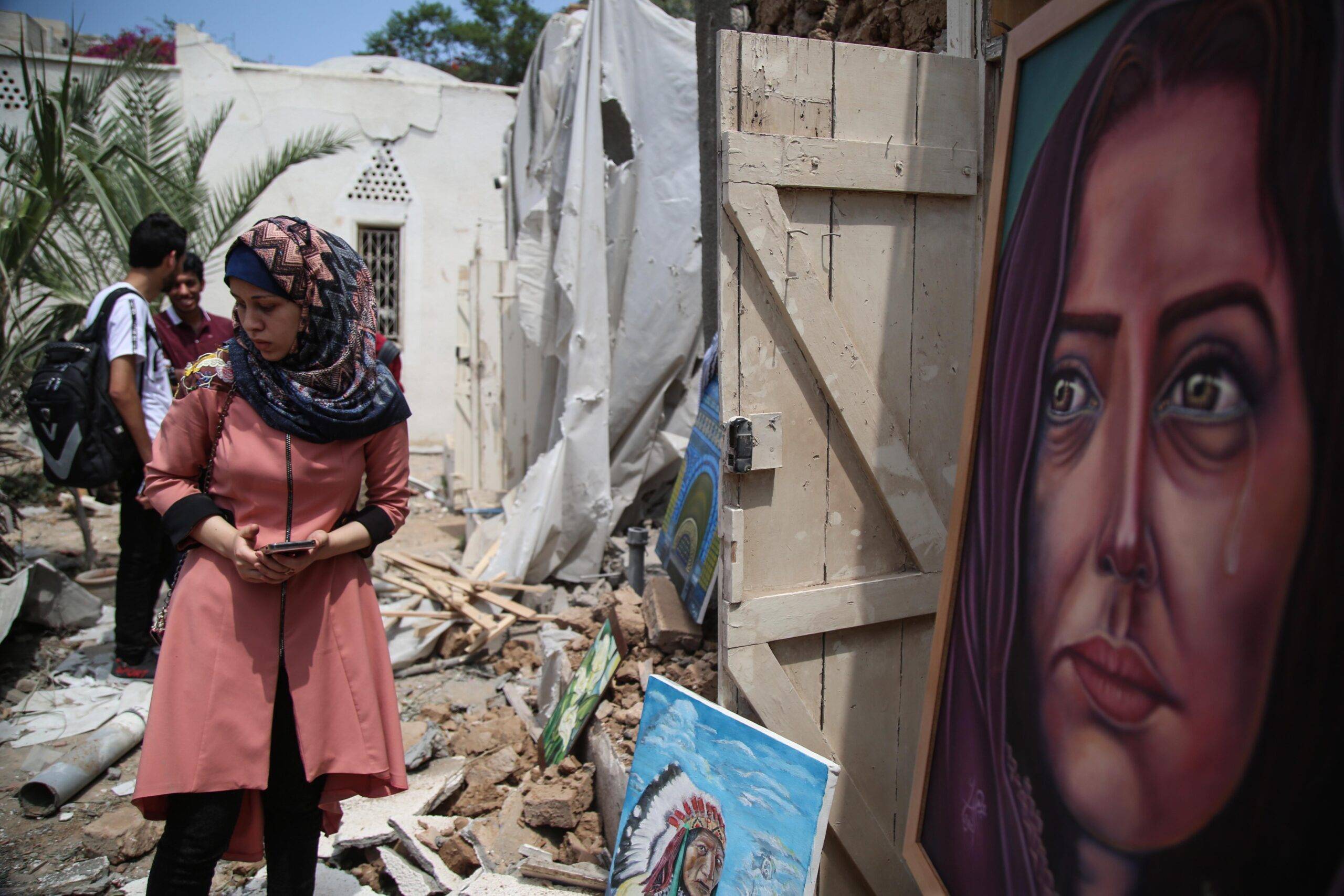 People take a look at paintings among wreckage of buildings during an exhibition that held by a group of Palestinian artists, after Israeli forces hit the area with airstrikes at the Art and Craft Village in Gaza City, Gaza on 16 July, 2018 [Hassan Jedi/Anadolu Agency]