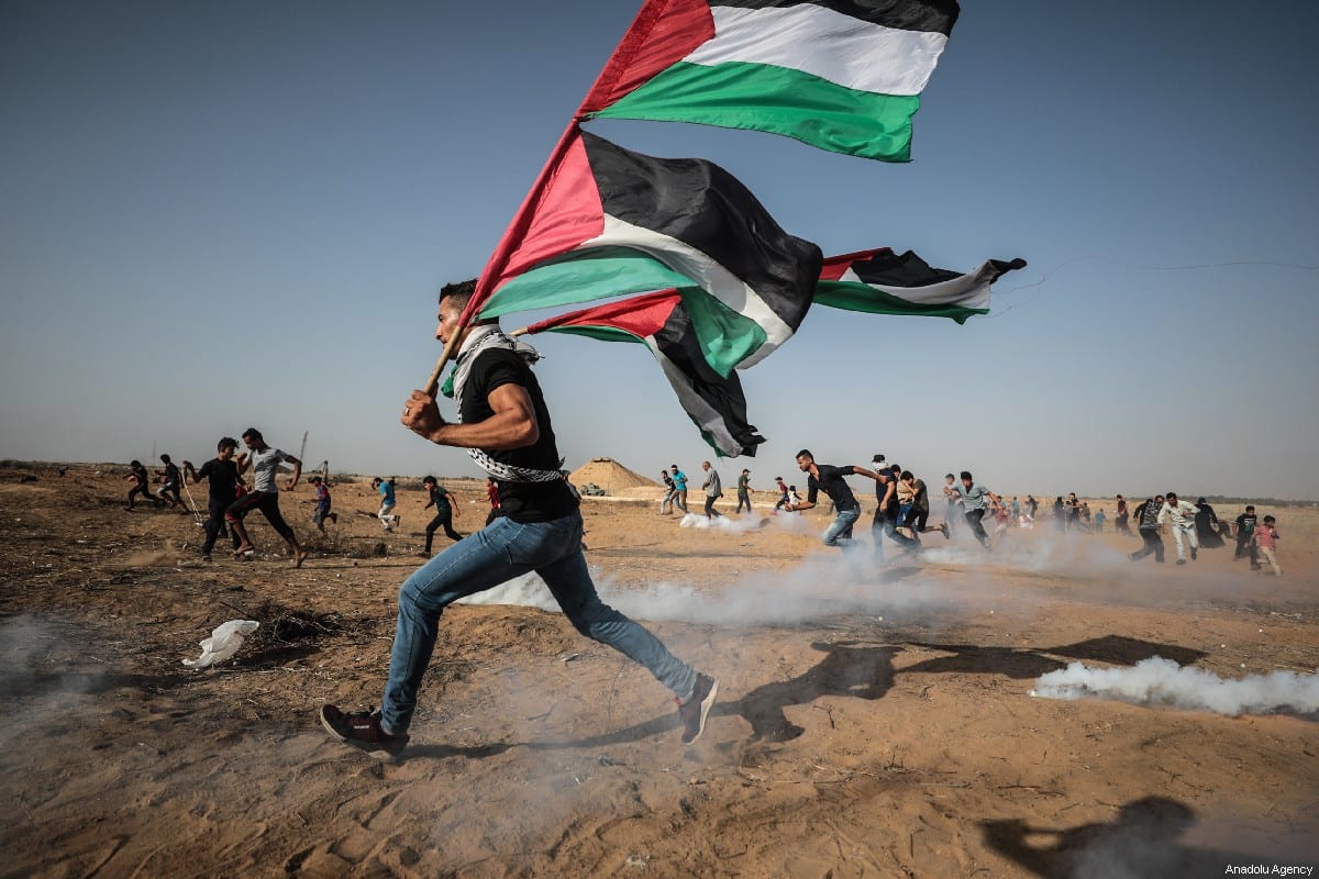 A protestor runs with the Palestinian flag, as Israeli forces fire tear gas during a demonstration during the "Great March of Return" at Israel-Gaza border in east of Khan Yunis, Gaza on July 12, 2019 [Mustafa Hassona / Anadolu Agency]