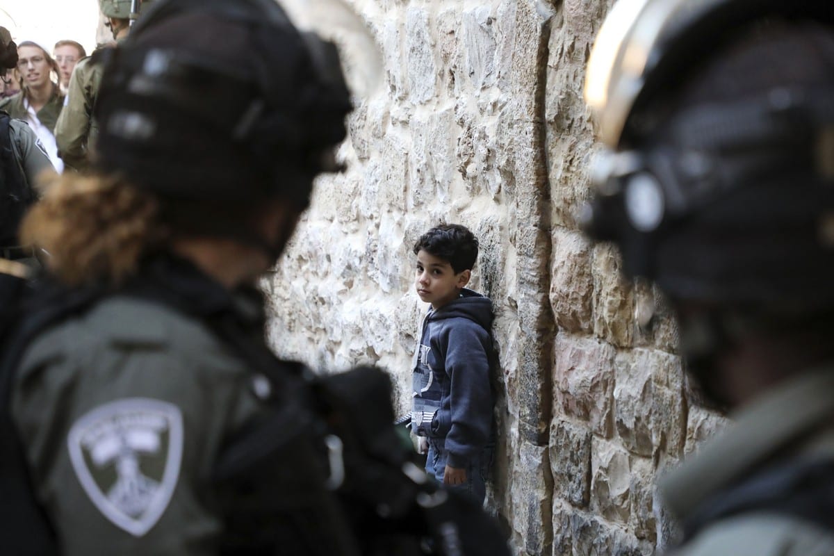 Israeli soldiers look at a Palestinian boy as he waits by the wall for Israeli settlers touring the old city and market of Hebron in the occupied West Bank to pass, on 21 December 2019. [HAZEM BADER/AFP via Getty Images]
