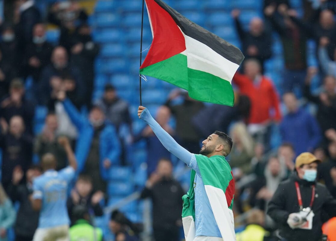 Riyad Mahrez of Manchester City wears the flag of Algeria, as he carries the flag of Palestine after Manchester City are presented with the Premier League Trophy following their victory in the Premier League match between Manchester City and Everton at Etihad Stadium on 23 May 2021 in Manchester, England. [Dave Thompson - Pool/Getty Images]