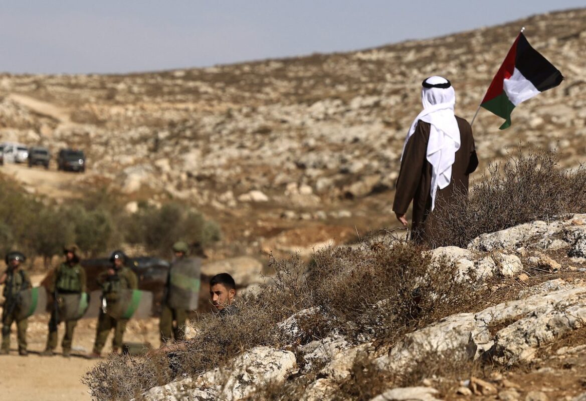 A Palestinian protester raises a national flag as he stands facing Israeli security forces, during a demonstration against the establishment of Israeli outposts on Palestinian lands, in Beit Dajan, east of Nablus in the occupied West Bank, on 12 November 2021. [JAAFAR ASHTIYEH/AFP via Getty Images]