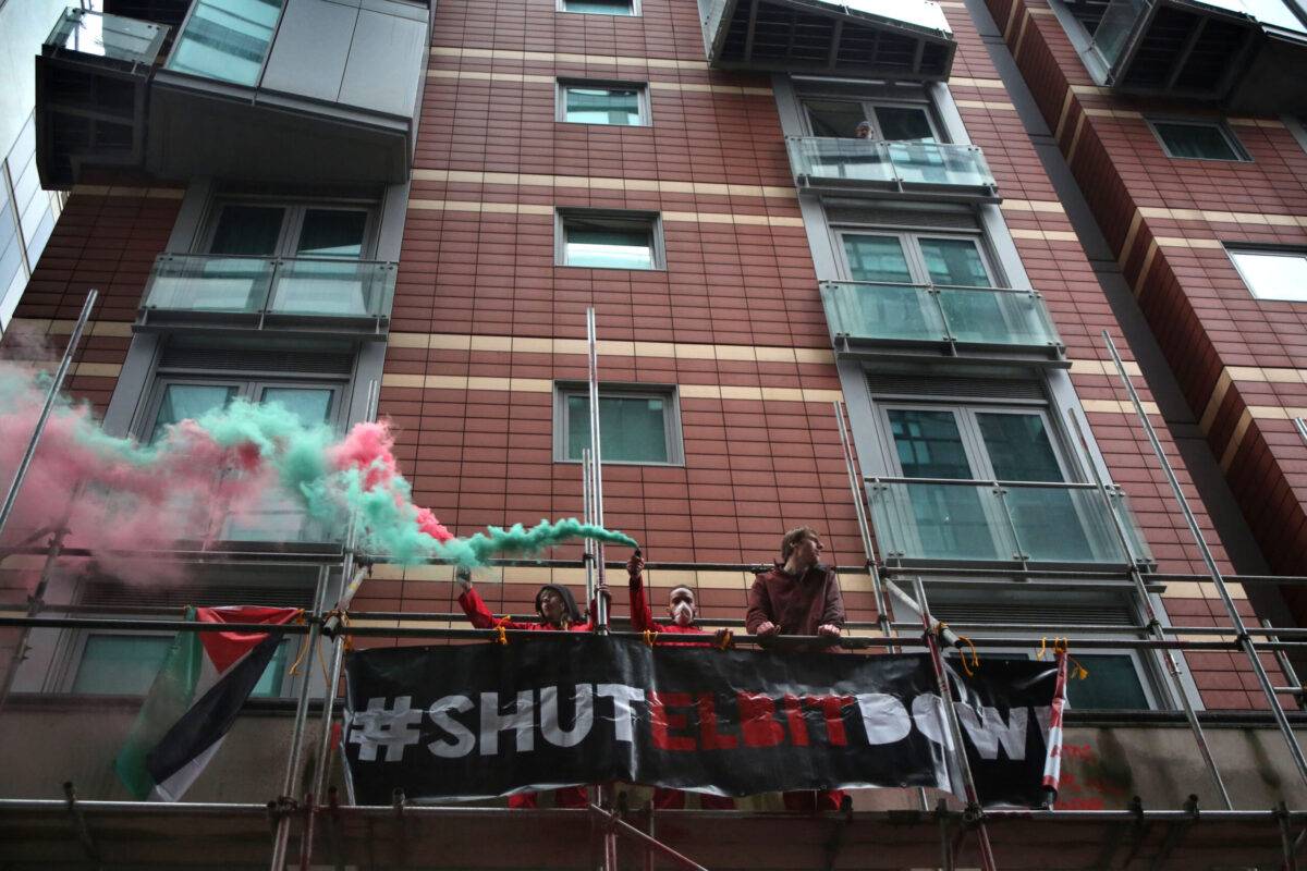 Activists from Palestine Action let off green and red smoke flares as they occupy scaffolding outside the offices of Aerospace Defence Security (ADS) on the Albert Embankment, watched by a resident from above, on March 28, 2023 in London, England [Martin Pope/Getty Images]