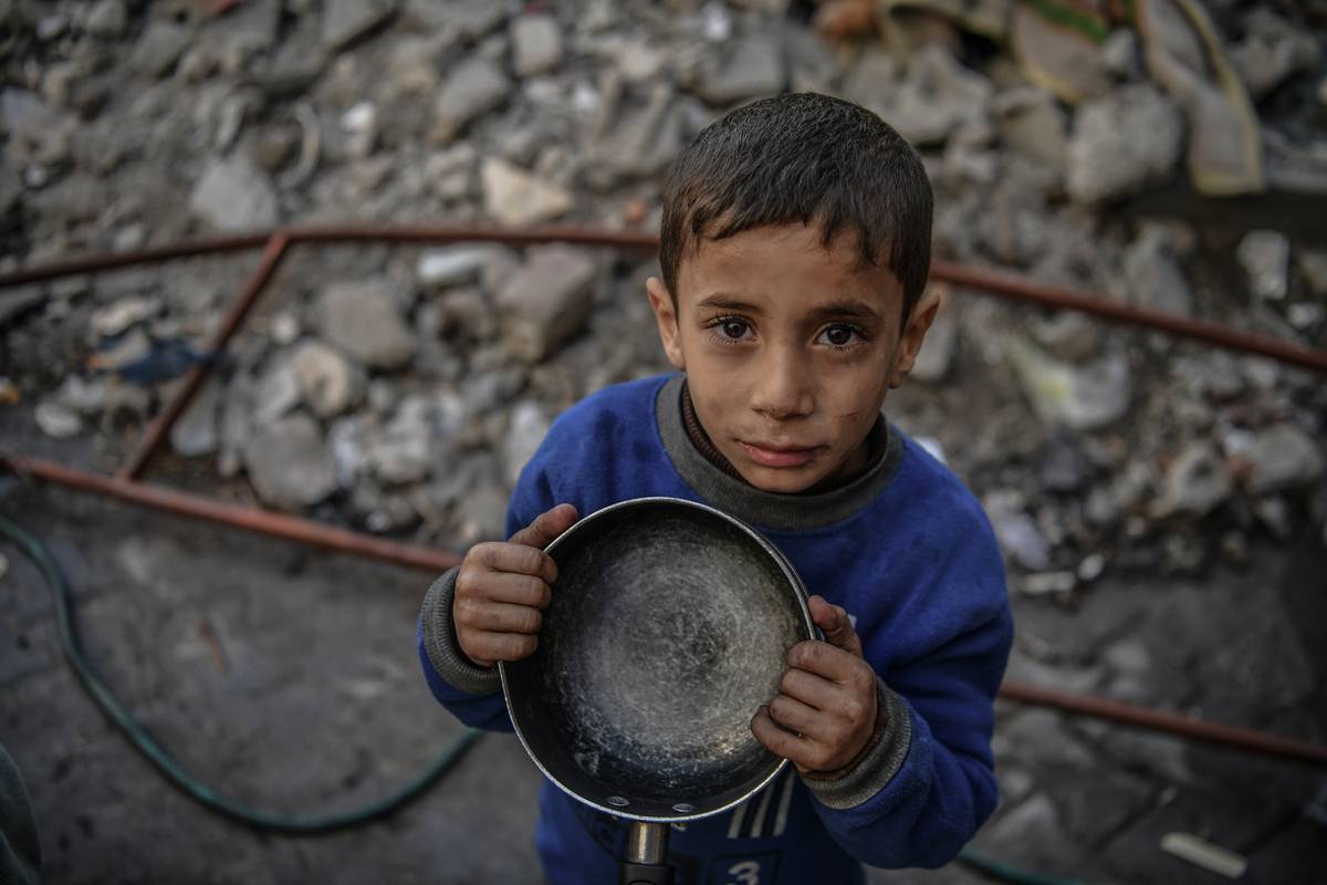 A Palestinian child holds an empty pot as he waits to receive food distributed by volunteers for Palestinian families displaced by Israeli attacks in Rafah, Gaza on 22 December, 2023. [Abed Zagout – Anadolu Agency]