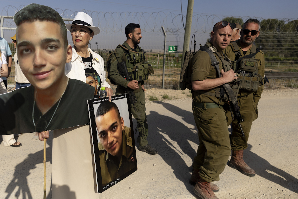 Varda Ben Baruch holds photos of her grandson Israeli- American hostage Edan Alexander as she walks near Israeli soldiers during a rally of families of hostages held in the Gaza Strip at Nir Oz near the border with the Gaza Strip on April 20, 2025 in Nir Oz, Israel. [Amir Levy/Getty Images]
